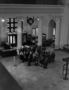 A school group meets its guide-lecturer. Class of children or teenagers at north entrance, Stanley Field Hall. Includes clock, The Book Shop and to the right postcards of Malvina Hoffman sculptures. Taken from 2nd floor balcony. For Raymond Foundation.