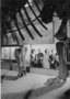 Two women and two male adult visitors looking at Brontosaurus excelsus, now Apatosaurus, skeleton, Fossil Vertebrates, Hall 38 exhibit.  Posed. Charles Knight mural of T. rex and Triceratops in background